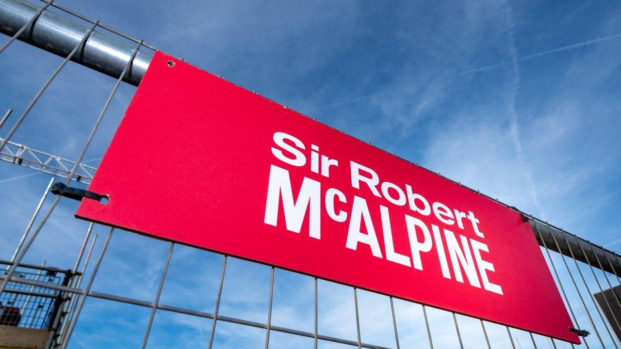 A red Sir Robert McAlpine sign attached to a metal construction fence against a blue sky