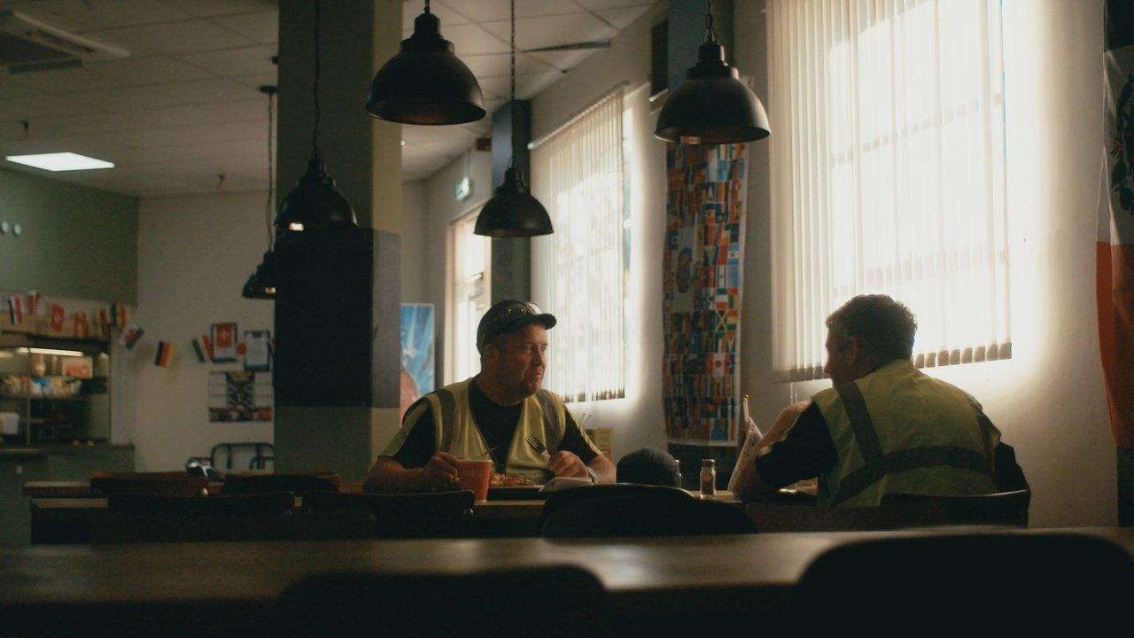 Two construction workers in high-visibility vests sit at a café table beneath hanging lamps.