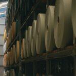 Large rolls of material stored on tall warehouse shelves beside a narrow aisle.