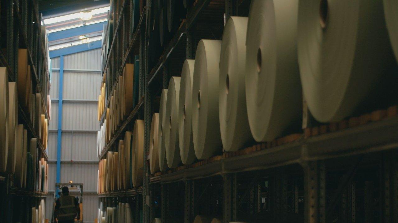 Large rolls of material stored on tall warehouse shelves beside a narrow aisle.