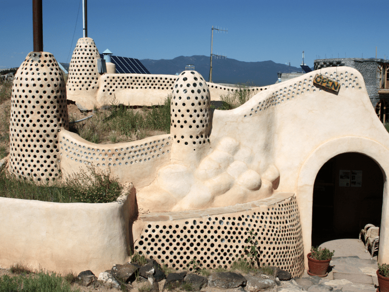 An earthship visitor centre with curved adobe walls studded with glass bottle ends and solar panels on the roof, mountains behind