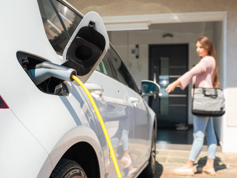 A white electric car charging on a driveway with a yellow cable while a woman walks towards the front door