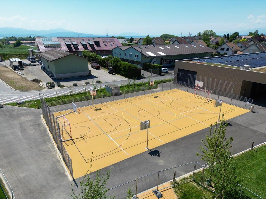 An aerial view of an outdoor multi-sport court with yellow surfacing, basketball hoops, and soccer goals, surrounded by buildings and roads, with a lake and mountains in the background under a clear sky.
