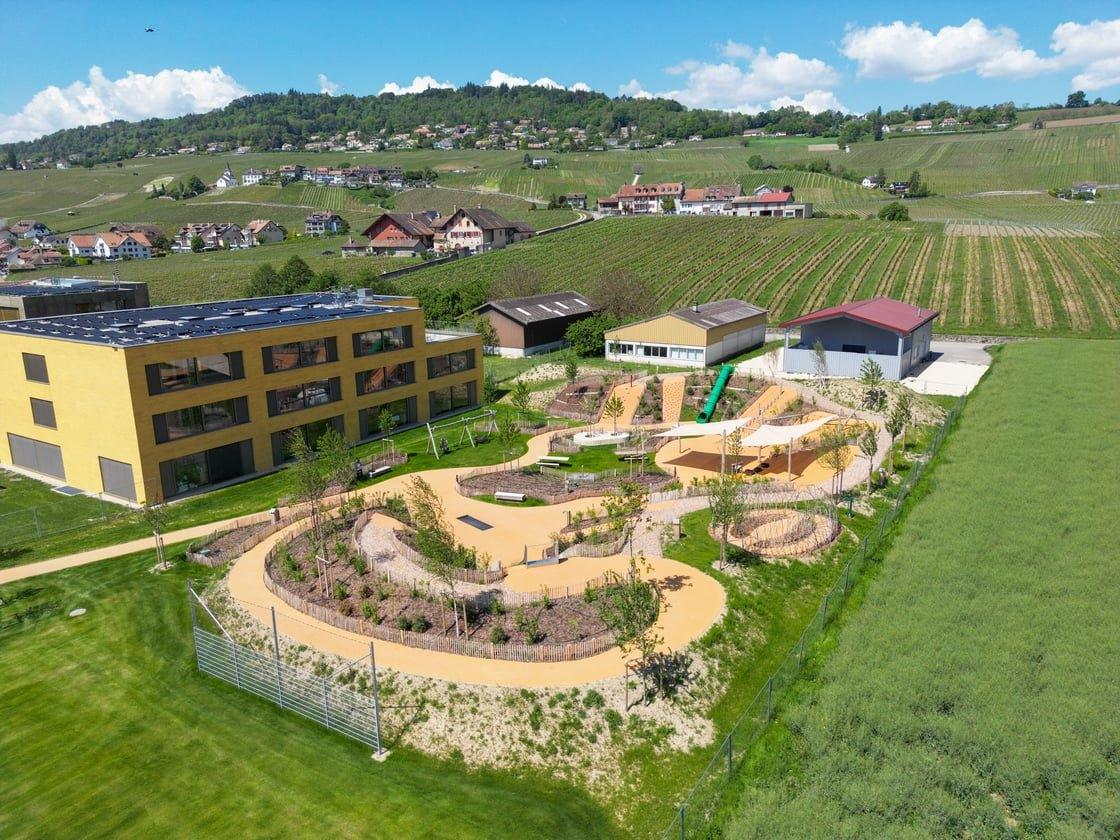 An aerial view of a modern yellow building next to a playground with vineyards and houses on rolling hills in the background.