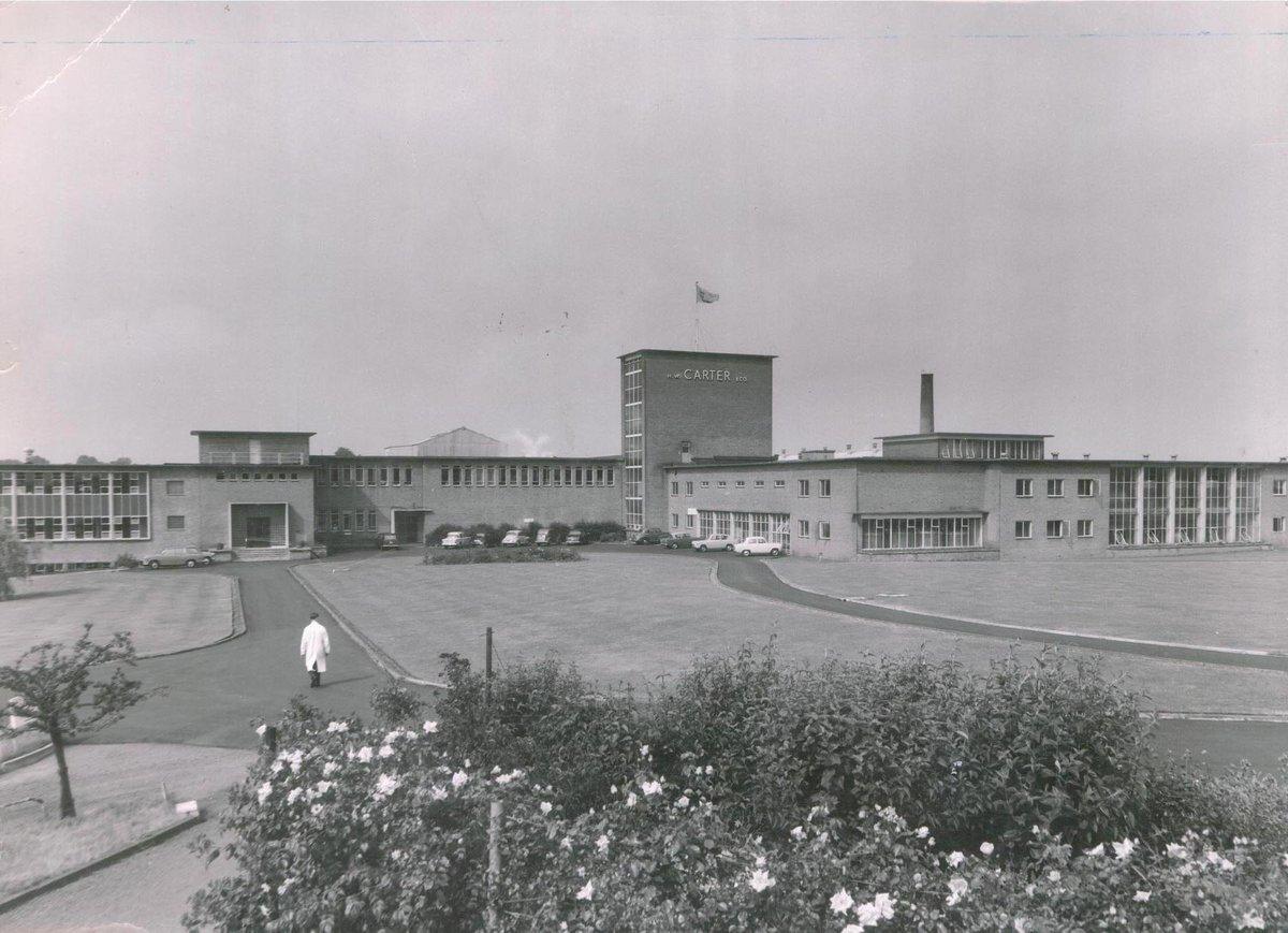 A black and white photograph of a large Carrier factory facility with a person in a lab coat walking on a path in the foreground.