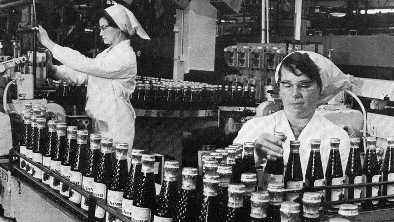 Two women in lab coats and headscarves work on a factory production line, handling bottles of a dark liquid.