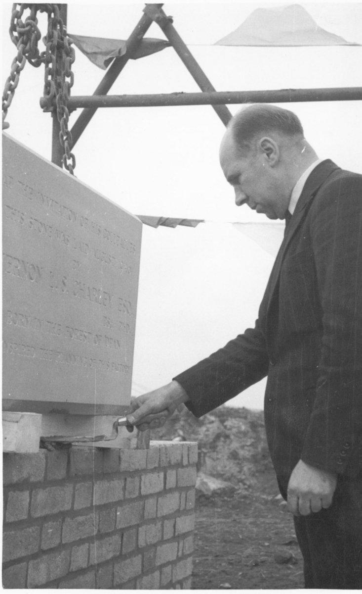 A man in a suit uses a trowel to lay a foundation stone on a brick wall, with scaffolding and a construction site visible in the background.