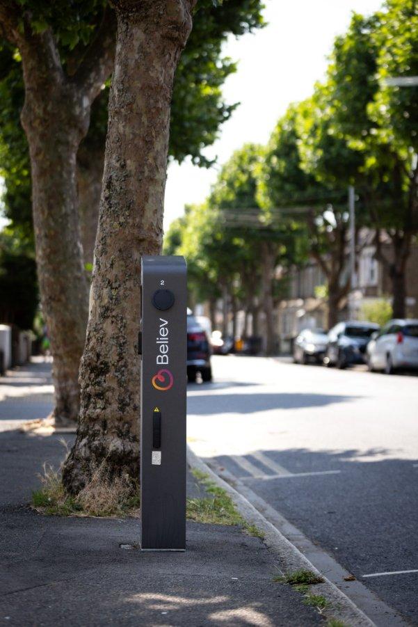 An electric vehicle charging station with the "Believ" logo on a sidewalk next to a tree-lined street with parked cars.