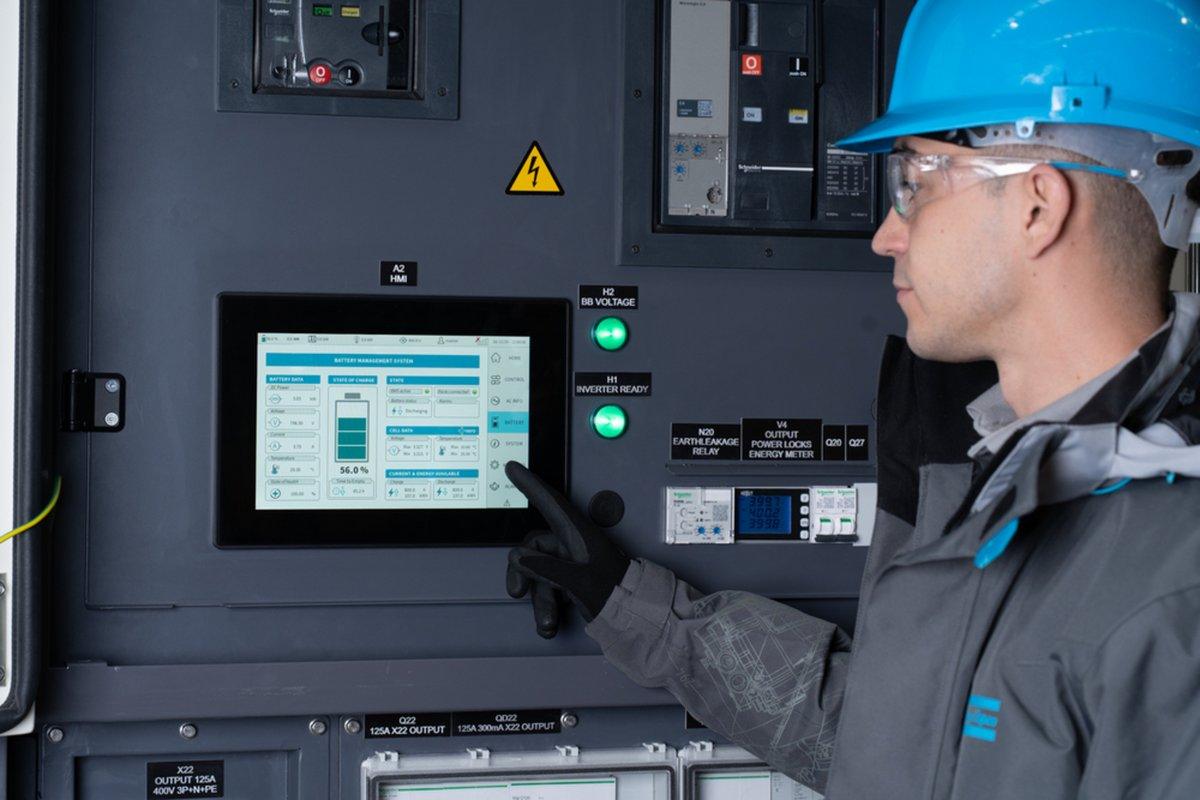 An engineer wearing a hard hat and safety glasses is touching a touchscreen display on an industrial control panel that shows a battery management system interface.