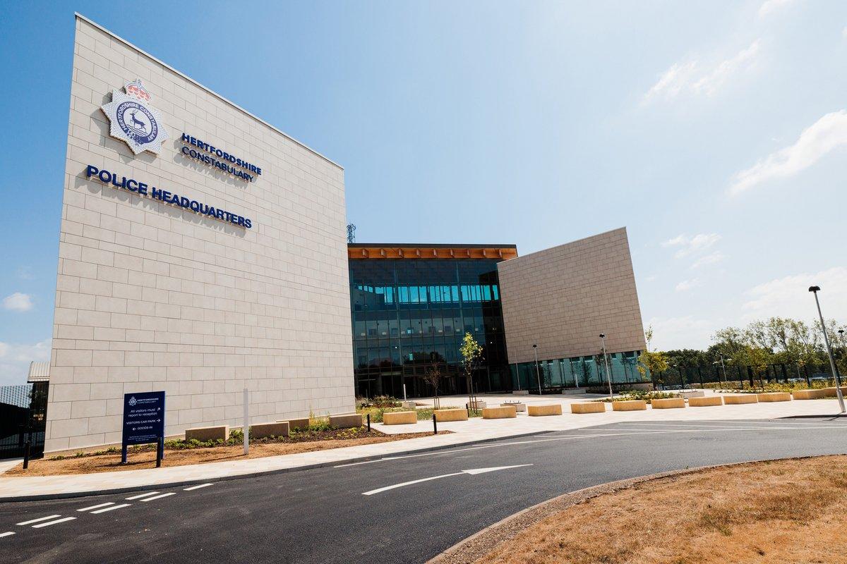 A modern, light-colored police headquarters building with the Hertfordshire Constabulary logo and name, and large glass sections, under a clear blue sky.