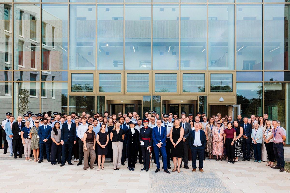A large group of people, some in uniform, stand posing for a photo outdoors in front of a modern glass building.