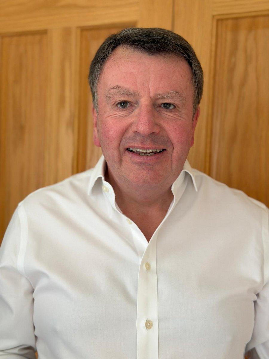A man in a white collared shirt smiles at the camera, standing in front of a wooden background.