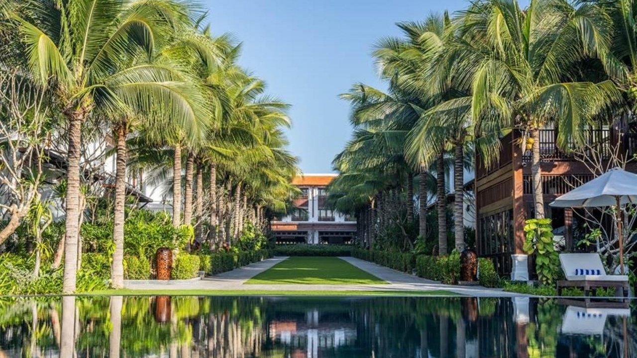 A tranquil resort scene featuring a long swimming pool reflecting palm trees and a central grassy path leading towards a large building in the distance under a clear blue sky.