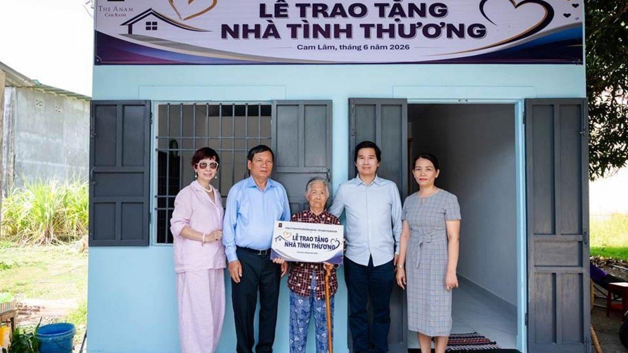 Five people, including an elderly woman holding a sign, stand in front of a light blue house with a banner on its facade.