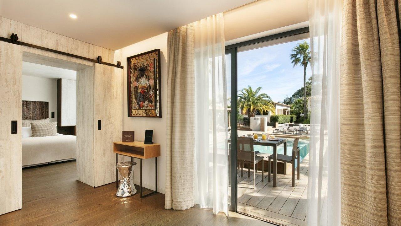 A bright hotel suite or room featuring a large sliding glass door opening to an outdoor patio with a swimming pool, palm trees, and an outdoor dining set. Inside, a console table with a stool and a framed artwork are visible, along with a partial view of a bedroom through open sliding barn doors.