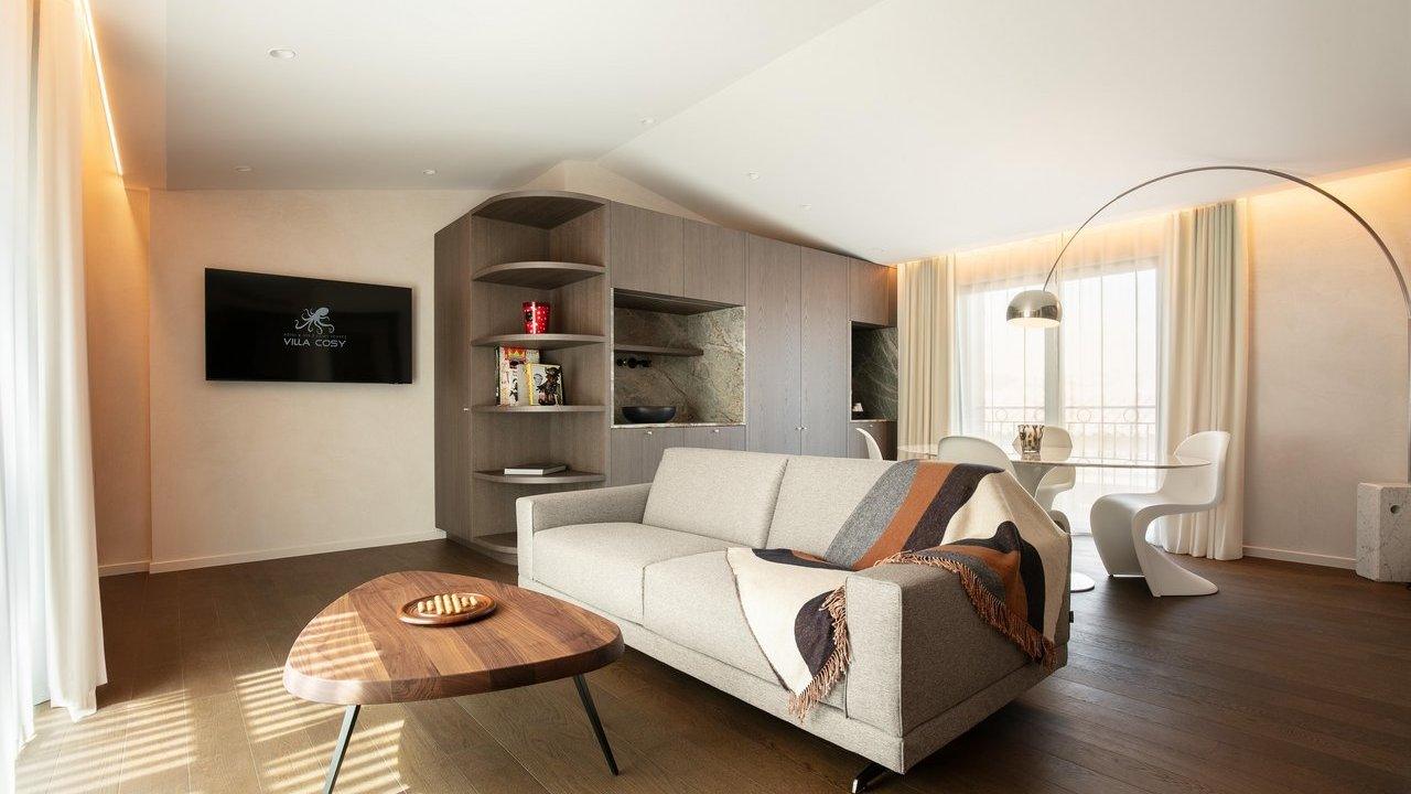 A modern, well-lit living room and dining area featuring a grey sofa with a throw blanket, a triangular wooden coffee table, built-in wooden shelving, a flat-screen TV, and a dining table with unique white chairs.