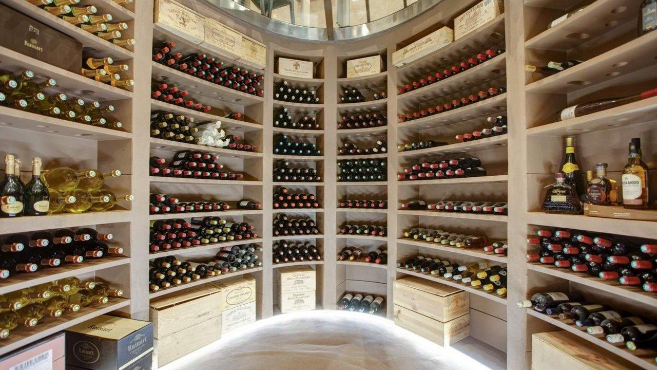 A well-lit, curved wine cellar with multiple shelves filled with various wine and liquor bottles, with wooden crates visible at the base.