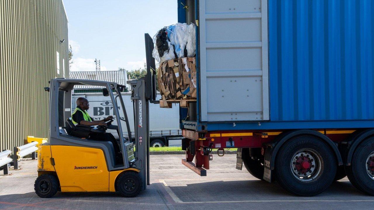 A man in a high-visibility vest operates a yellow forklift, loading bales of cardboard and plastic into a blue shipping container on a truck in an industrial setting.