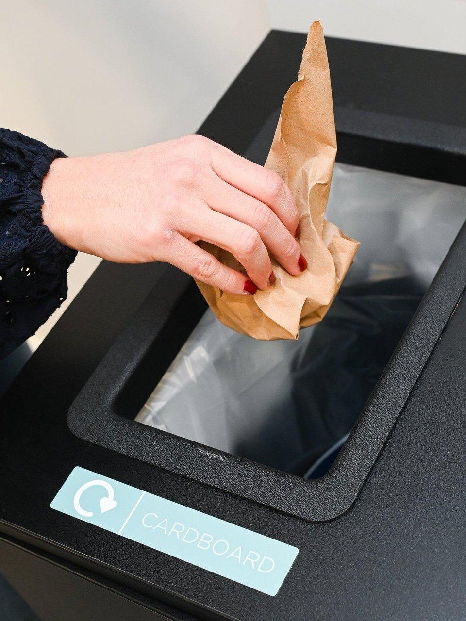 A person's hand places a crumpled brown paper bag into a black recycling bin labeled "CARDBOARD" with a recycling symbol.