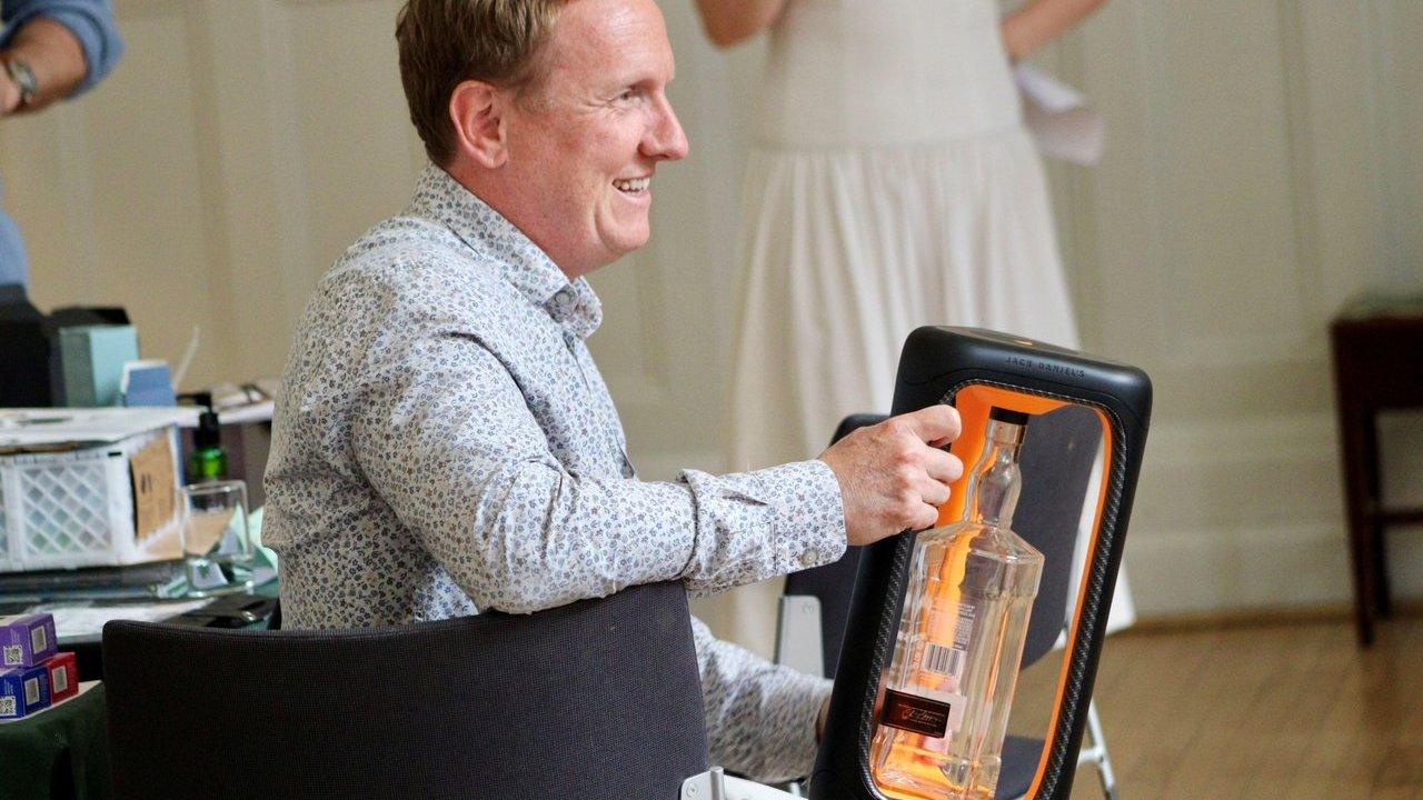 A man in a patterned shirt smiles while holding a clear bottle in an orange and black display case, with an indoor background.