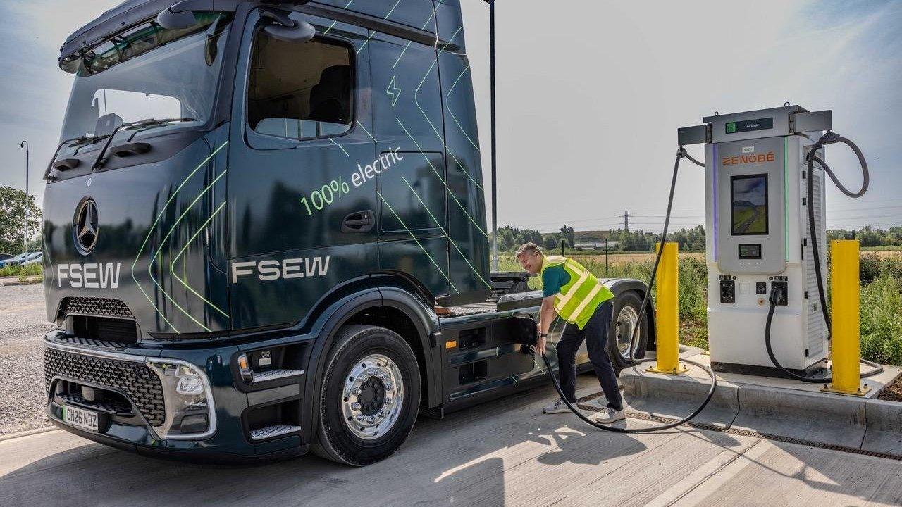 A person in a high-visibility vest is charging a dark green Mercedes-Benz electric truck at an outdoor Zenobe charging station.