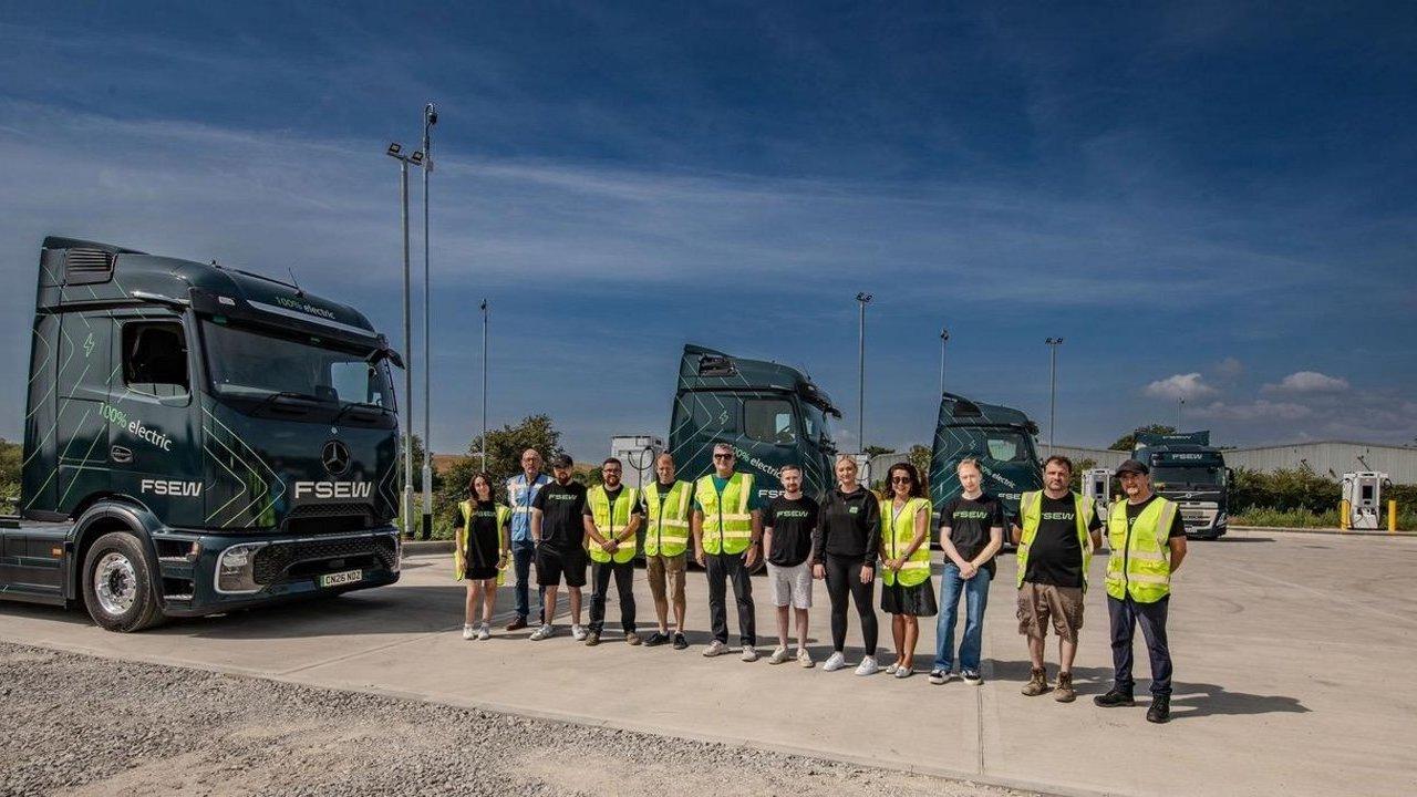 A group of people stand in front of multiple dark green electric Mercedes-Benz trucks at an outdoor charging station under a blue sky.