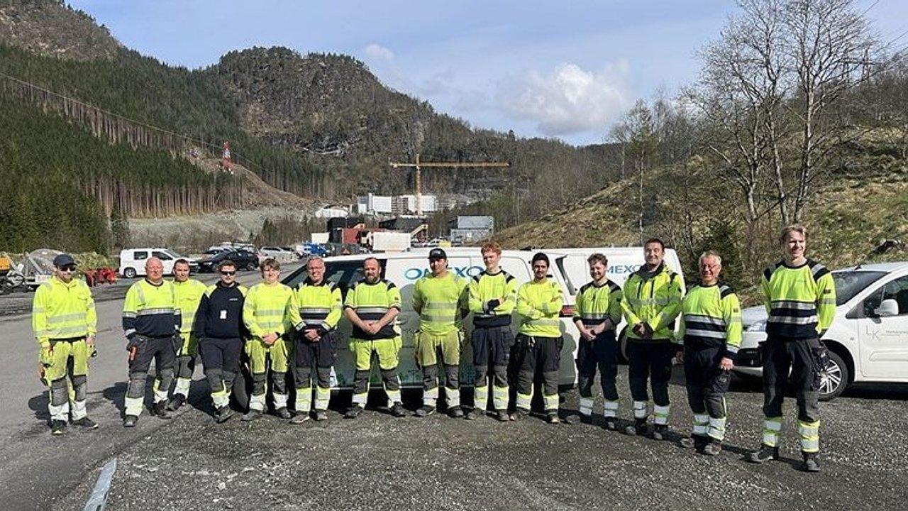 Eleven construction workers in high-visibility clothing stand beside work vehicles at a mountain site.