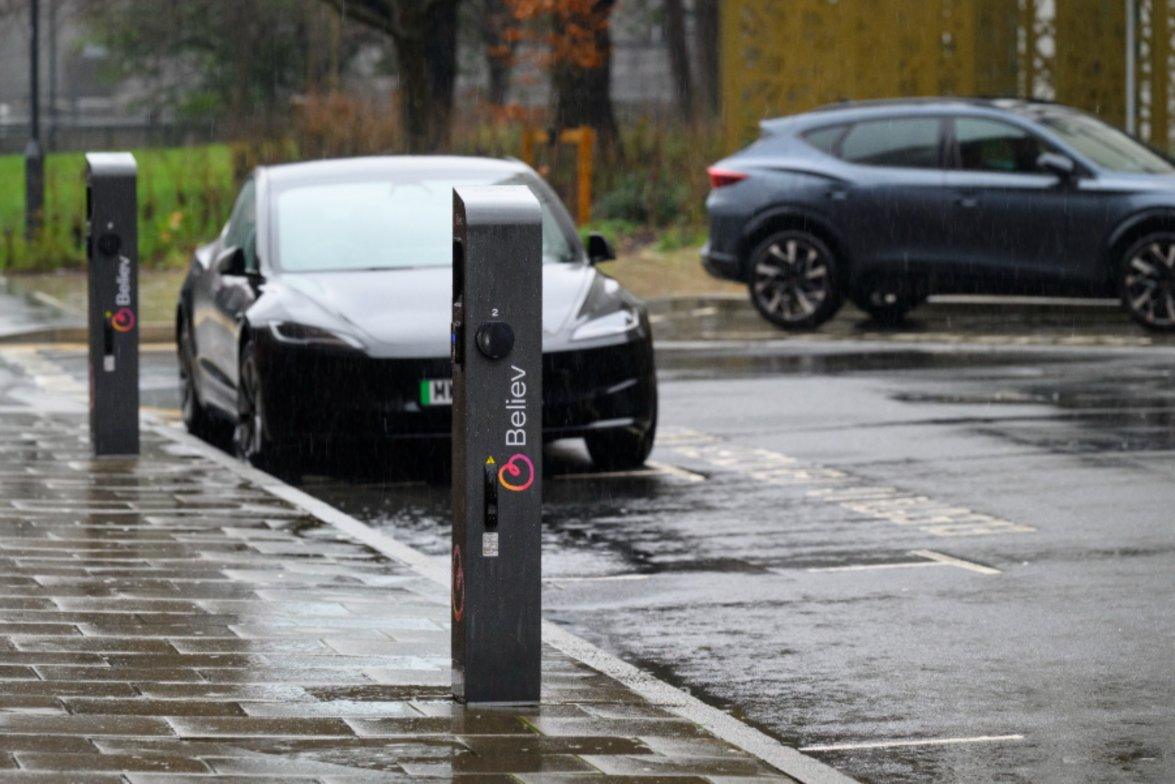 Two Believ electric vehicle charging stations on a rainy street with cars parked nearby.