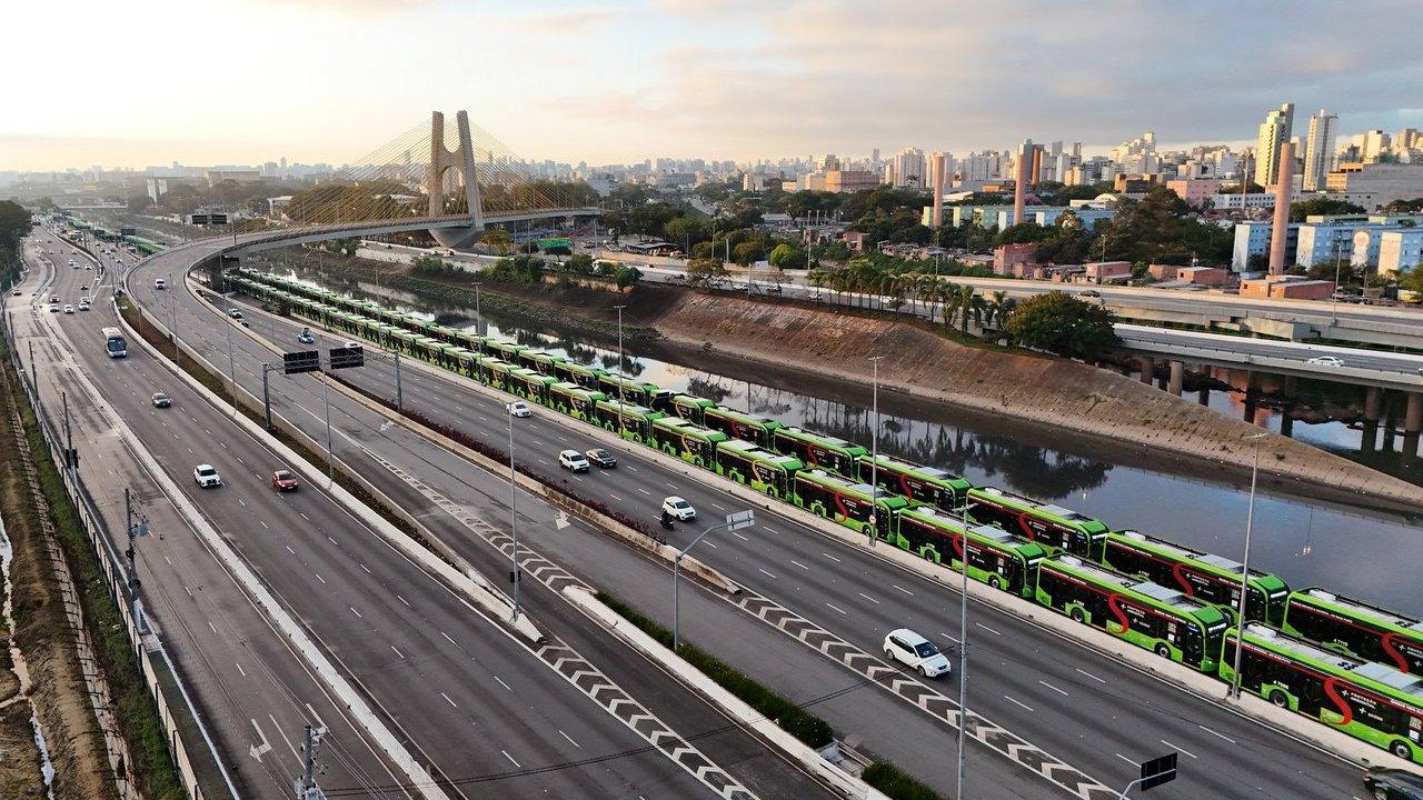 Aerial view of a long line of green electric buses on a highway with a city skyline and cable-stayed bridge behind