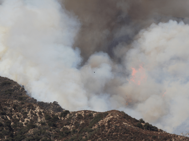 Thick smoke and orange flames rising from a wildfire burning across chaparral-covered hills