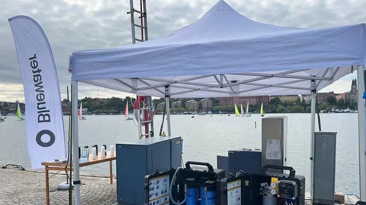 A white canopy with equipment beside a lakeside, with sailboats on the water and a vertical banner.