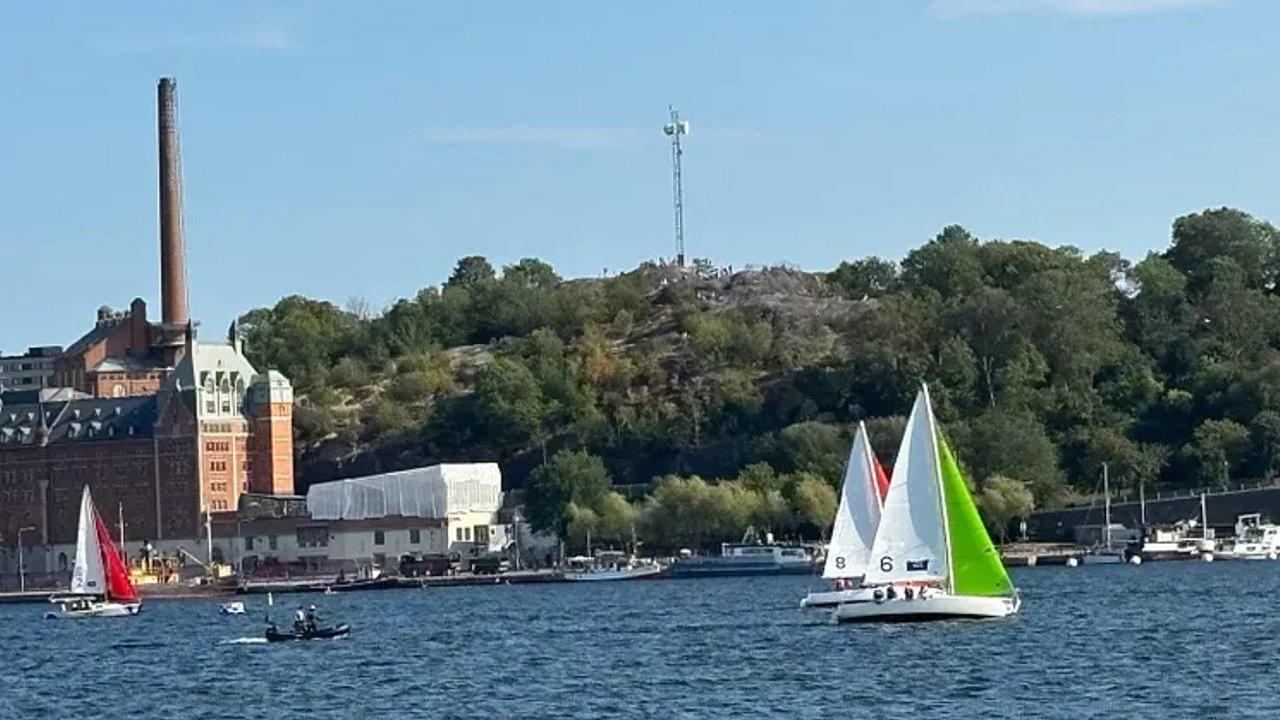 Sailboats with colorful sails on a harbor beside a brick industrial building and wooded hill.