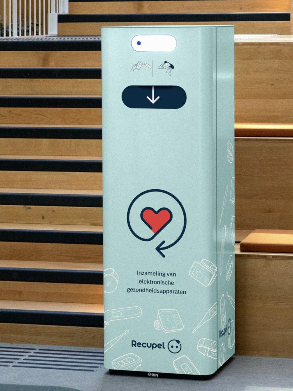 A Recupel collection bin for electronic health devices beside a wooden staircase.