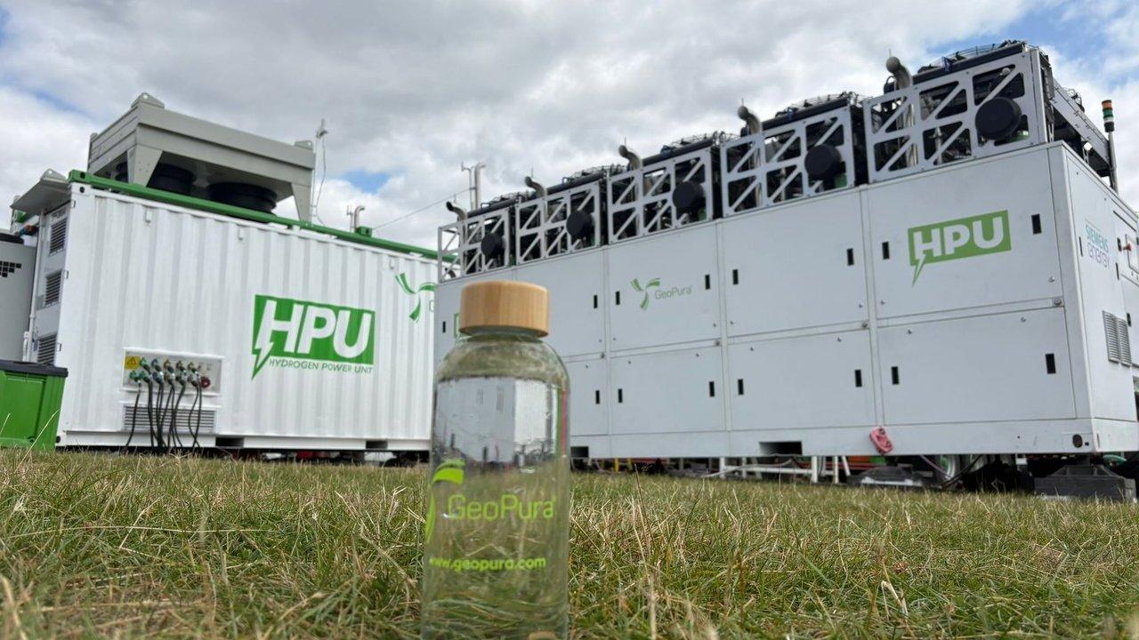 A GeoPura water bottle in grass before hydrogen power units under a cloudy sky.