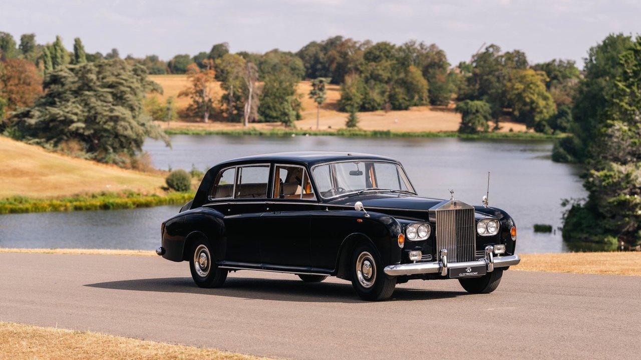 A black vintage luxury sedan parked beside a lake with trees and dry grass in the background