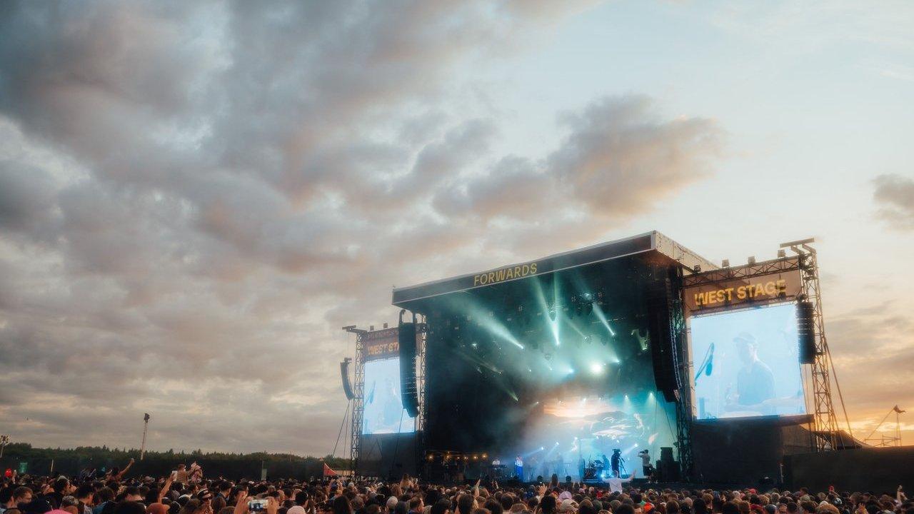 Crowd watching performers on an outdoor festival stage with bright lights at sunset