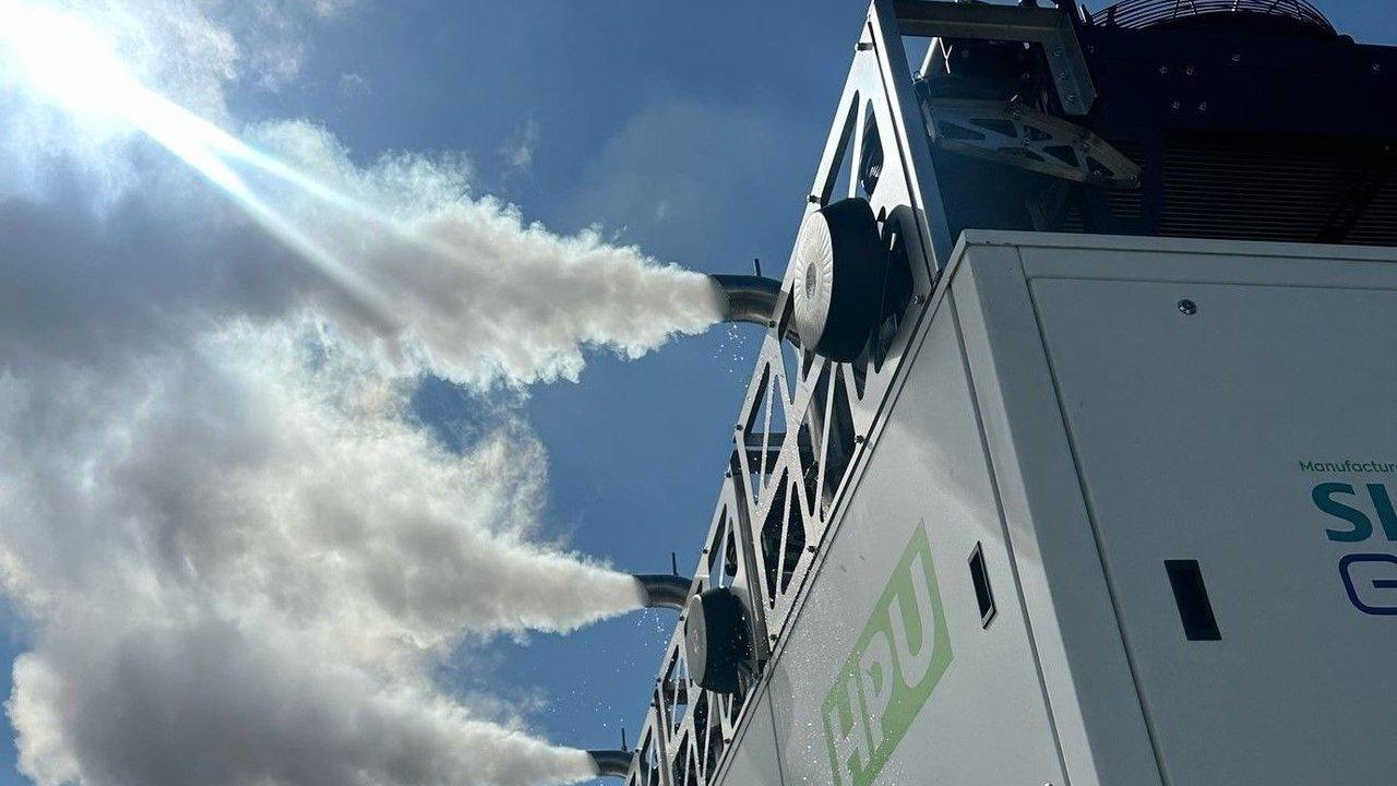 Industrial machine spraying thick white mist into a blue sky beneath bright sunlight