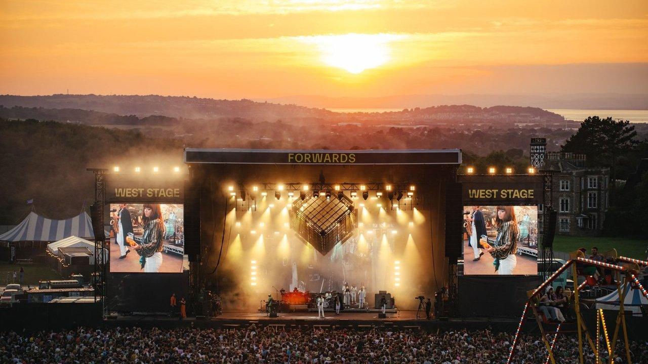 A crowded outdoor music festival stage lit at sunset, with large video screens and a fairground ride in the foreground