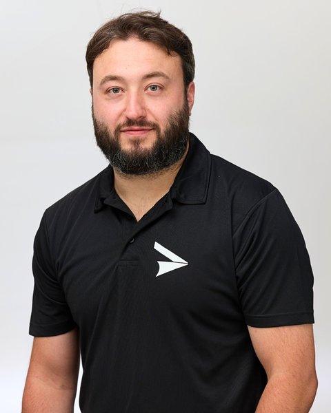 A bearded man wearing a black polo shirt with a white arrow logo against a light gray background
