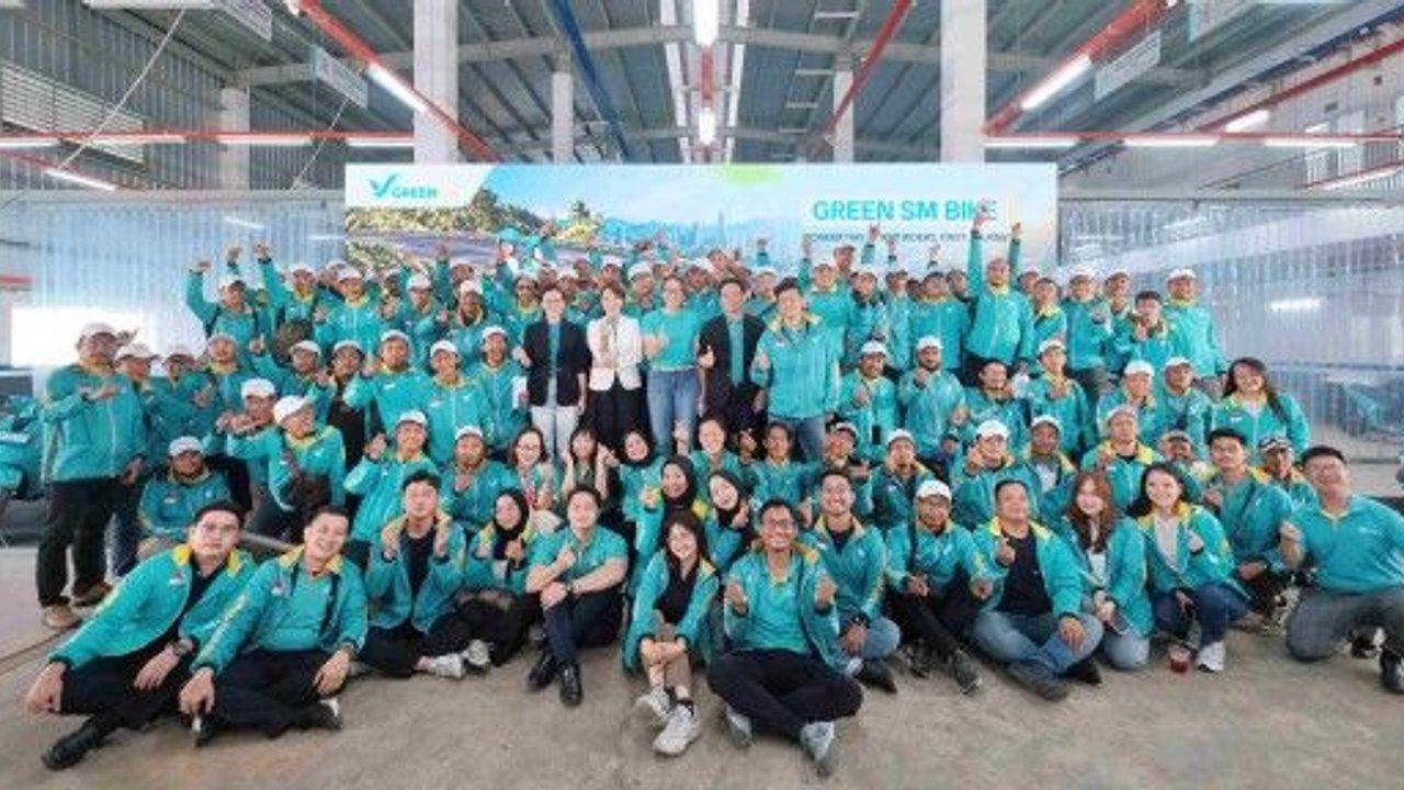Large group of workers in teal jackets posing inside a warehouse beneath a Green SM Bike banner