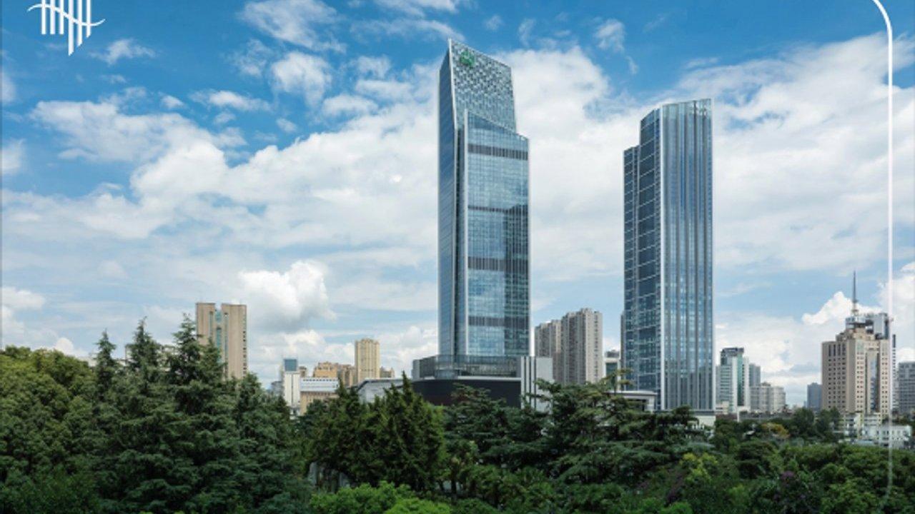 Two glass skyscrapers rising above trees and surrounding city buildings under a cloudy blue sky.