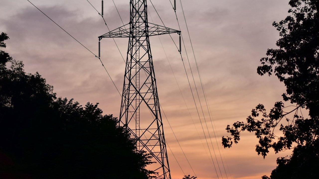 Electricity transmission tower and power lines silhouetted against a sunset sky, framed by trees.