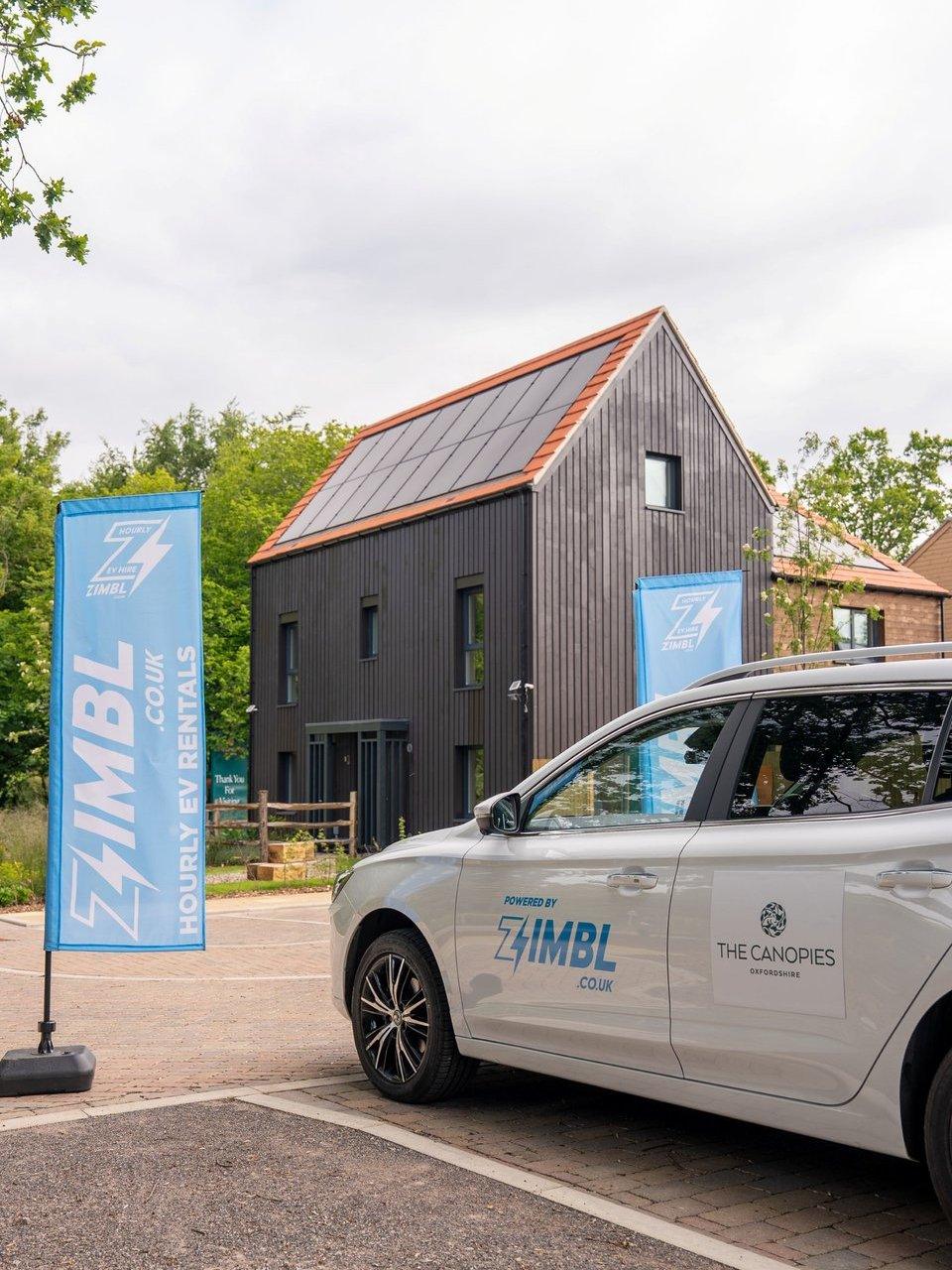 Zimbl-branded car and flags outside a black timber house with solar panels.