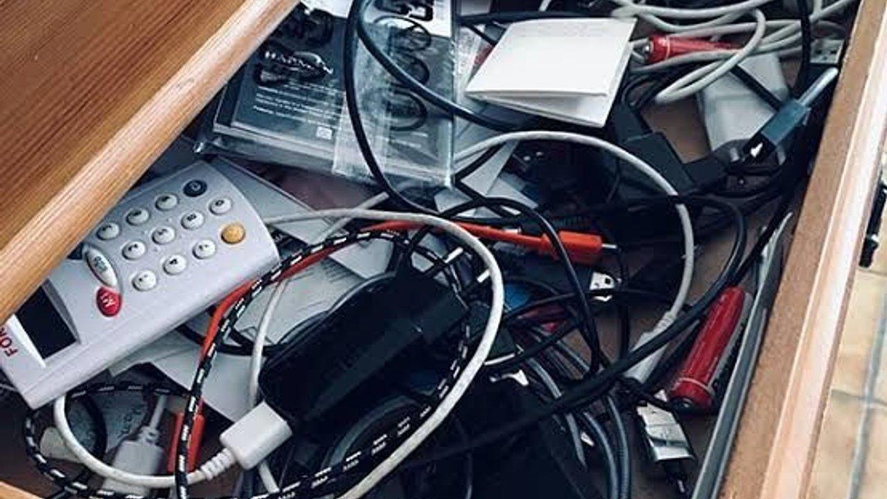 A wooden drawer filled with tangled cables, chargers, adapters and old electronic devices.