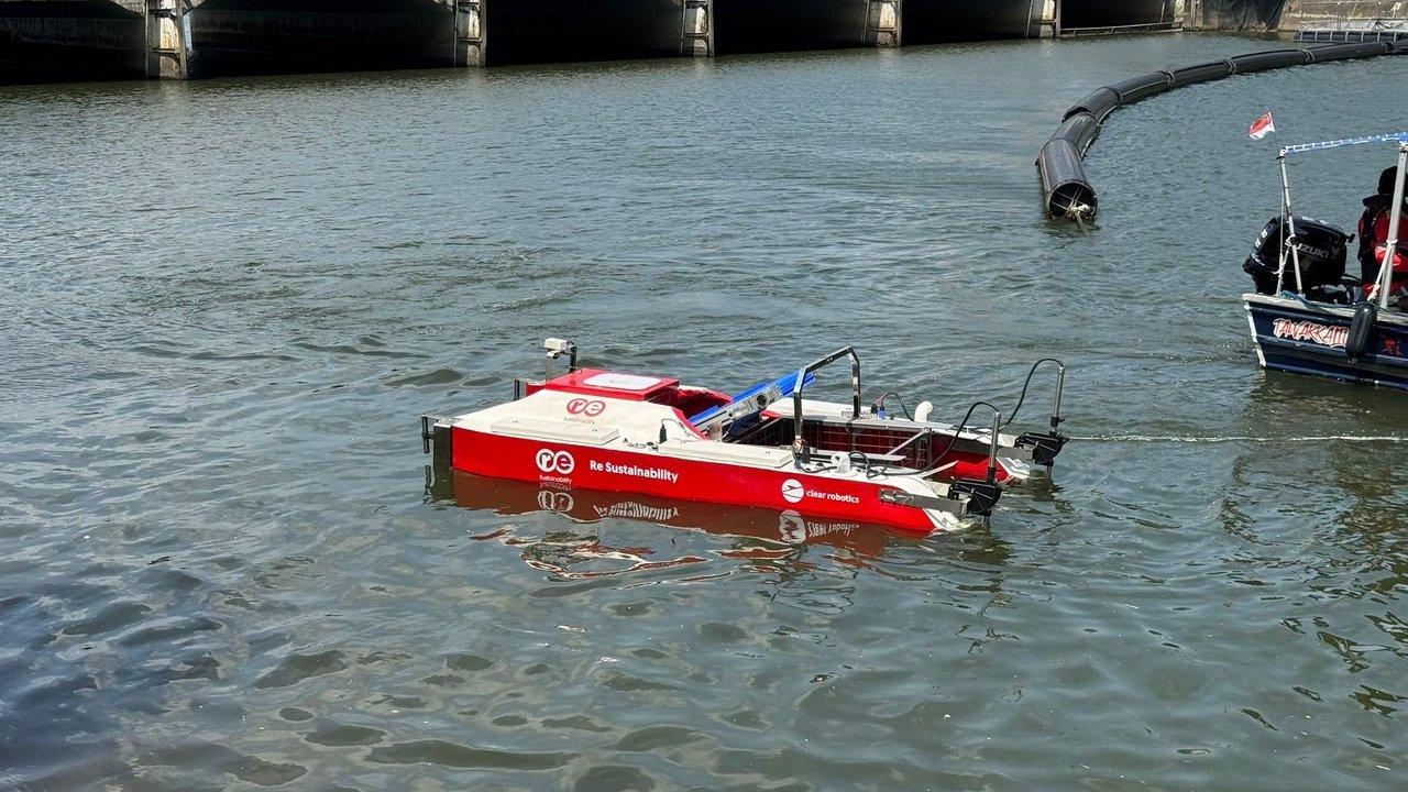 A red autonomous boat beside a floating barrier on a river, with another motorboat nearby.