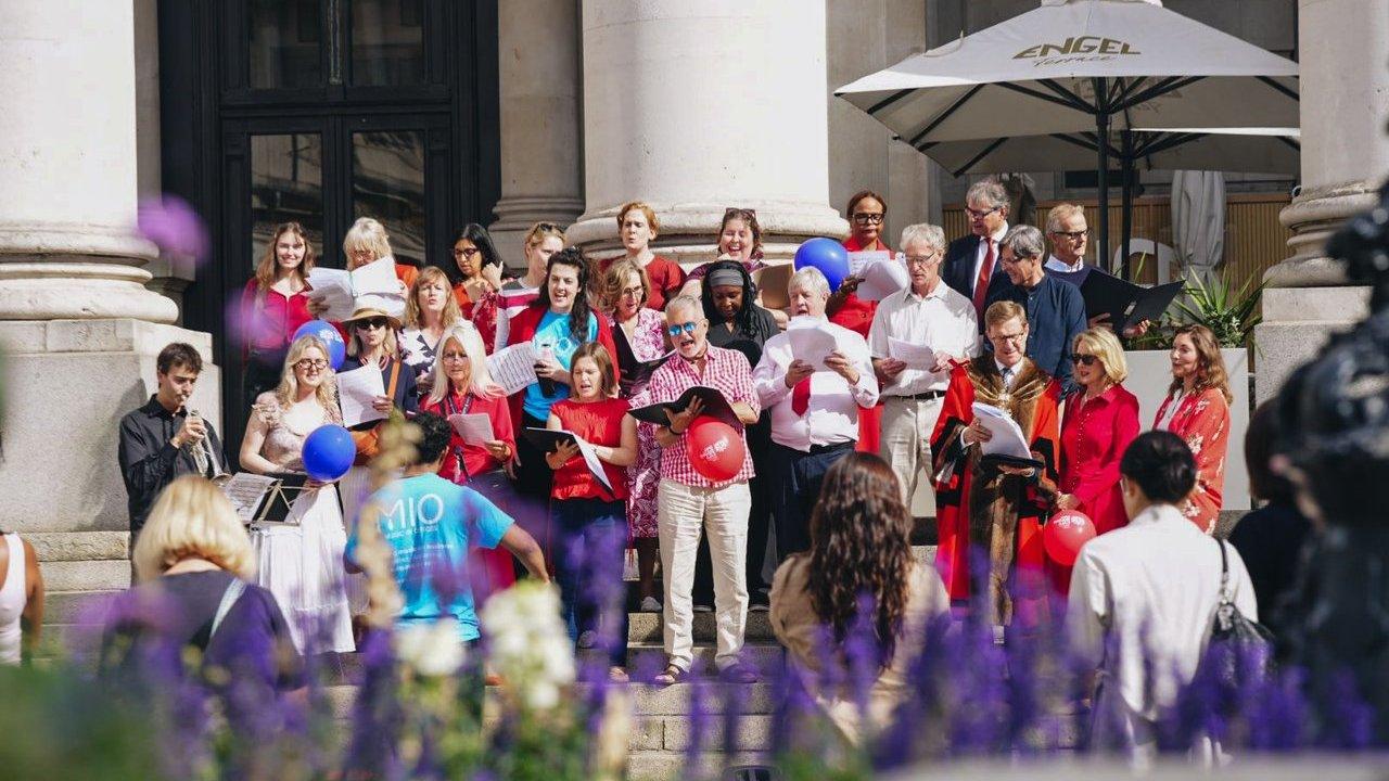 A choir sings on building steps as a trumpet player performs for an audience.