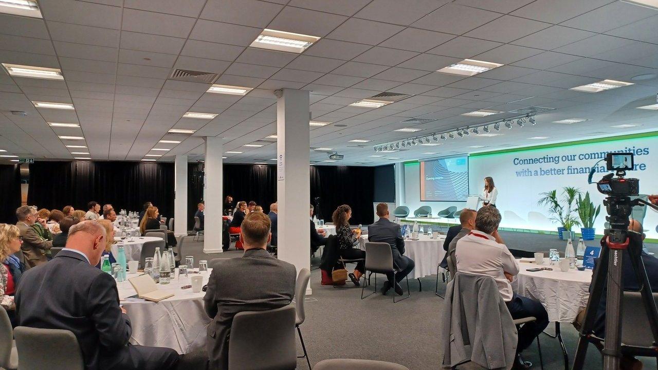 Audience seated at tables watches a woman speak beside a presentation screen and video camera.