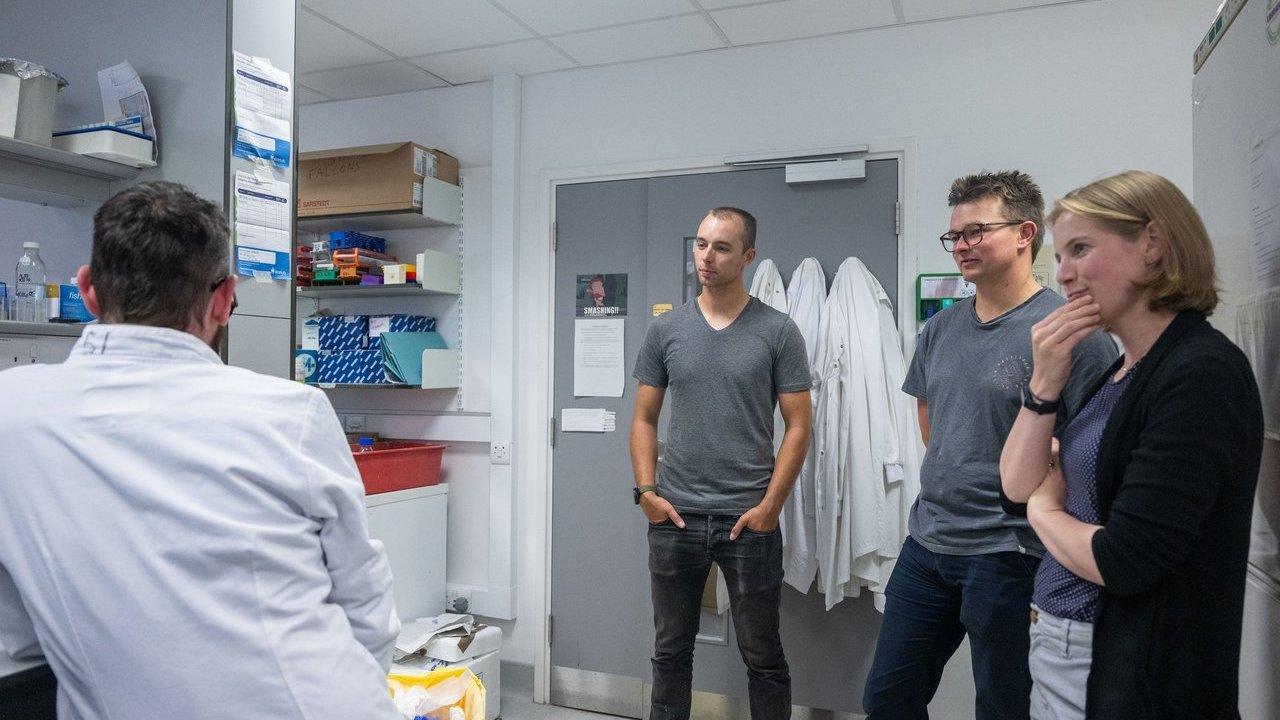 Three people observe a lab worker in a white coat in a laboratory.