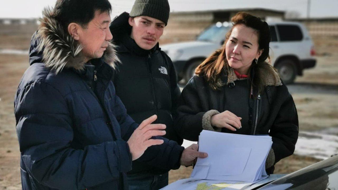 Three people in winter coats discuss papers beside a vehicle in a rural area.