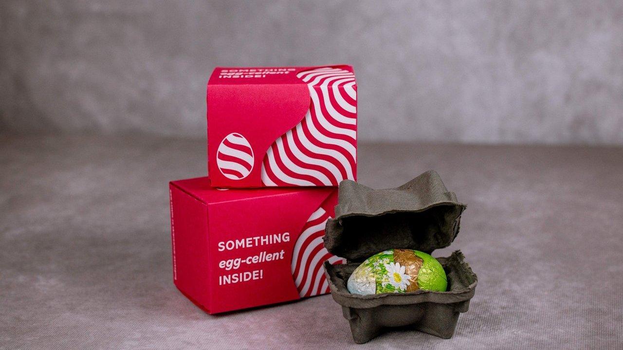 Two red patterned boxes beside an open egg carton holding a foil-wrapped chocolate egg.